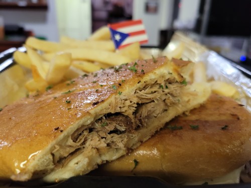 A close-up of a sandwich filled with pulled pork, served with fries and a small Puerto Rican flag.