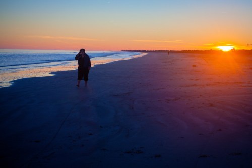 A person stands on a beach at sunset, with gentle waves and a colorful sky in the background.