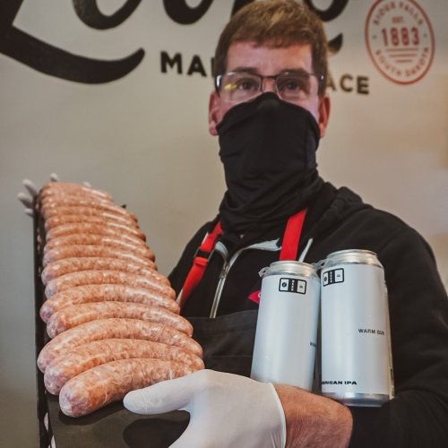 A masked man in gloves holds a tray of sausages and two cans of beer in a food market setting.