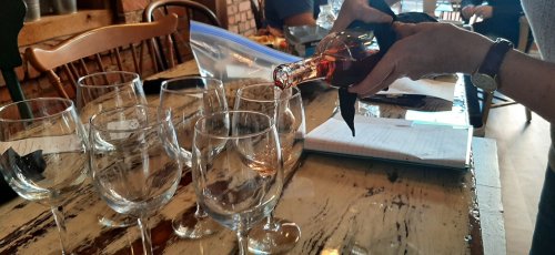 A hand pours a drink from a bottle into a wine glass, surrounded by empty glasses and a notebook on a rustic table.