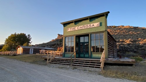 A green wooden building labeled "The Carissa" in a rustic setting with mountains in the background and a gravel path.