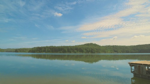 A serene lake scene with calm waters reflecting a blue sky and lush green hills in the background.