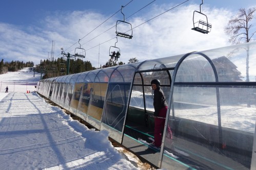 A person stands at the entrance of a covered ski lift, surrounded by snow and ski slopes in the background.