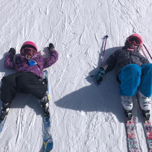 Two children in ski gear lying on the snow, wearing helmets and goggles, with skis beside them.