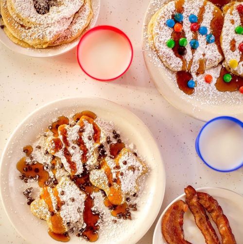 A colorful breakfast spread featuring pancakes, bacon, and milk in bowls, topped with chocolate chips and syrup.