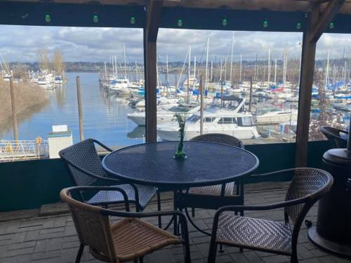 A waterfront view from a covered patio, featuring a table and chairs overlooking a marina with boats.