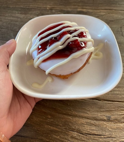 A hand holding a glazed donut topped with cherry filling and drizzled with white icing on a small plate.