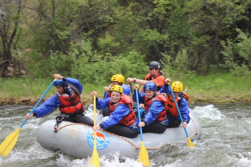 A group of six people in life jackets paddling a raft through a river, surrounded by greenery and splashing water.
