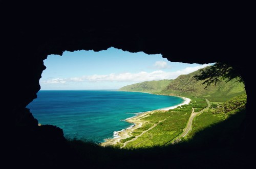 View of a coastal landscape framed by a cave opening, showcasing blue waters and green hills under a clear sky.