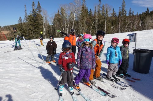 A group of children and adults in ski gear, smiling and posing on a snowy slope with trees in the background.