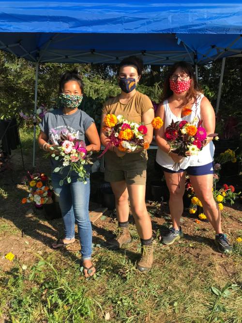 Three women wearing masks stand under a blue tent, holding colorful flower bouquets in a garden setting.
