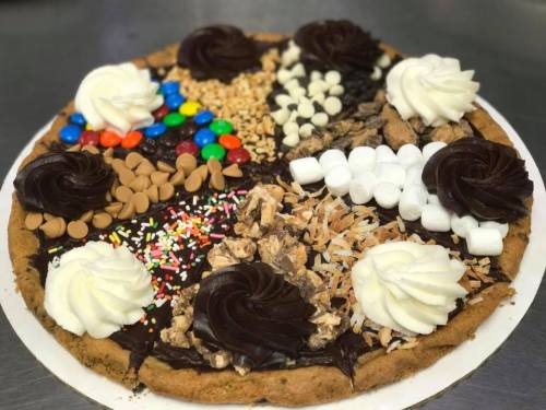 A large cookie pizza topped with chocolate frosting, whipped cream, nuts, candies, and marshmallows in colorful sections.