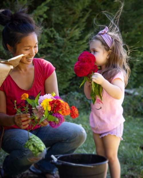 A woman and a young girl smile while arranging colorful flowers in a garden, enjoying a sunny day together.