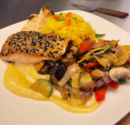 A plate featuring sesame-crusted salmon, yellow rice, sautéed vegetables, and a creamy sauce, garnished with herbs.