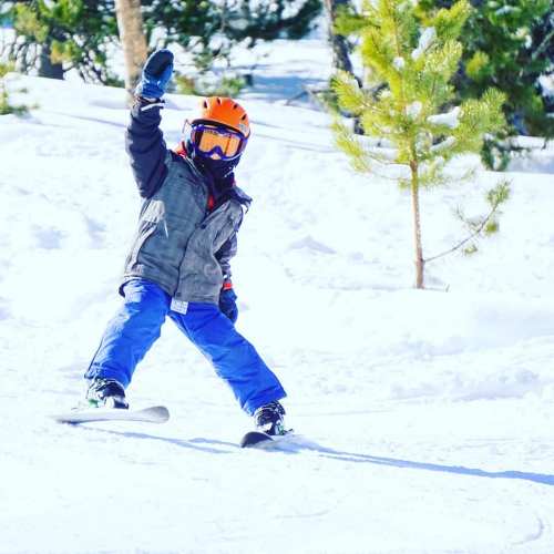 A child in a helmet and goggles snowboards on a snowy slope, raising one hand in excitement. Pine trees are in the background.