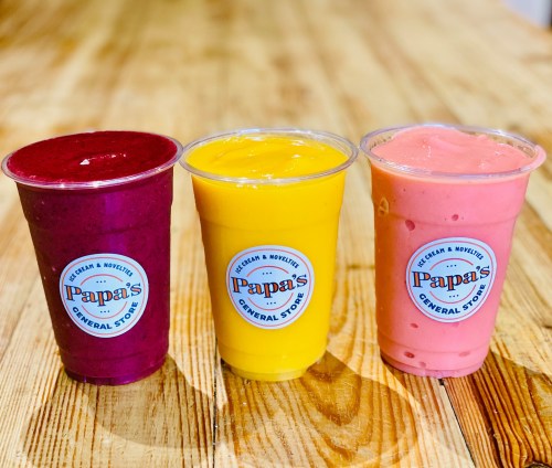 Three colorful smoothie cups from Papa's General Store: purple, yellow, and pink, on a wooden table.
