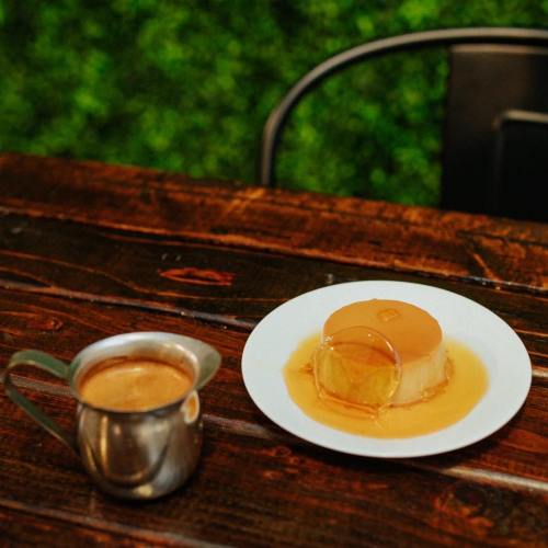A plate of flan with caramel sauce next to a small metal pitcher of coffee on a wooden table.