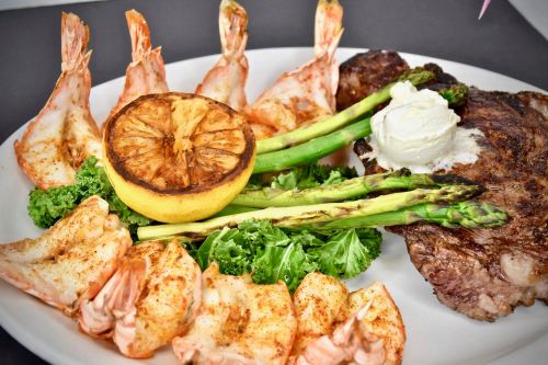 A plate featuring grilled shrimp, asparagus, a lemon half, and a steak, garnished with kale and a dollop of cream.
