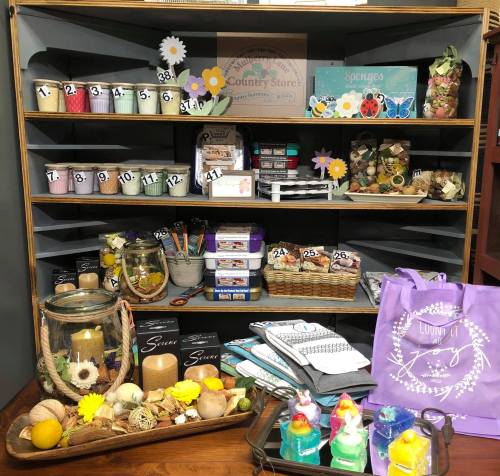 A display shelf filled with craft supplies, jars, sponges, and decorative items, alongside a tote bag and notebooks.