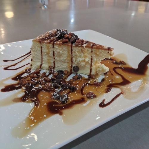 A slice of cheesecake topped with chocolate drizzle, crushed nuts, and a caramel sauce on a white plate.