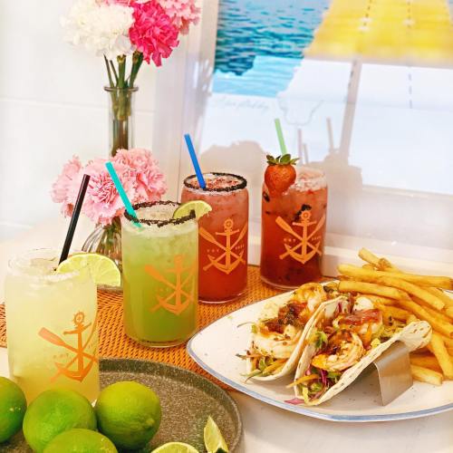 Colorful cocktails and tacos on a table, with fresh limes and fries, set against a beach-themed backdrop.