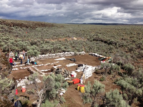 Archaeological dig site in a sagebrush landscape, with workers and equipment arranged in a grid pattern.