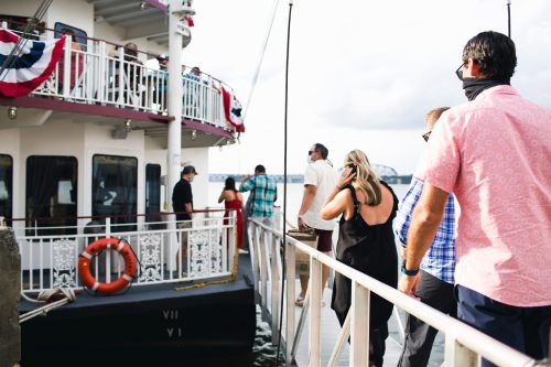 People wearing masks board a boat decorated with red, white, and blue, with a scenic waterfront in the background.