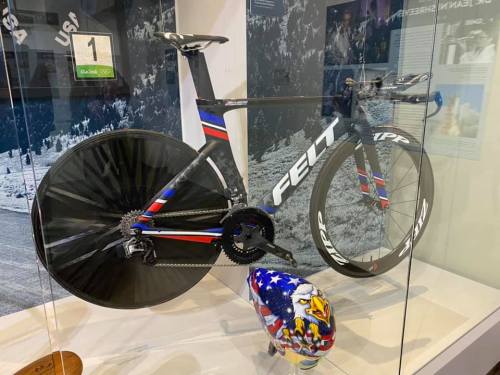 A sleek black racing bike displayed in a glass case, featuring a colorful design and an eagle-themed helmet beside it.