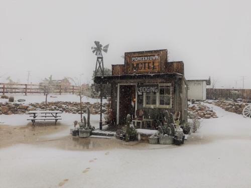 A snow-covered Pioneertown Motel with a windmill, surrounded by rocks and cacti, in a winter landscape.