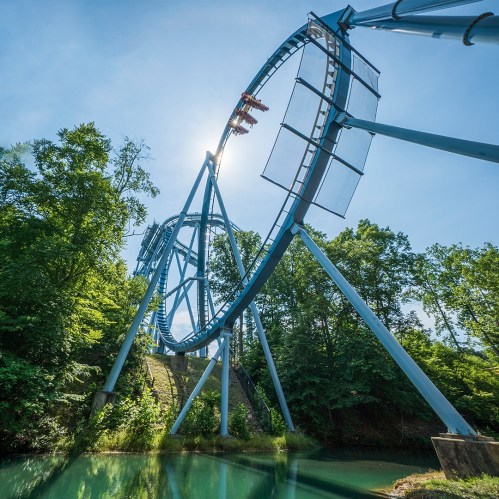 A blue roller coaster loop rises above a serene green landscape under a clear blue sky.