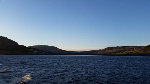 A serene view of a calm lake surrounded by rolling hills under a clear blue sky at sunset.