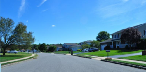 A quiet suburban street lined with houses, trees, and a clear blue sky.