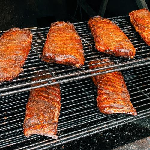 Close-up of several racks of smoked ribs on a grill, showcasing a rich, dark seasoning and a smoky texture.