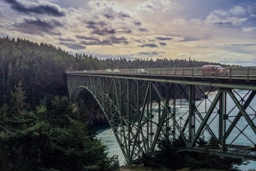 A green bridge spans a river, surrounded by trees and under a cloudy sky, with vehicles crossing.