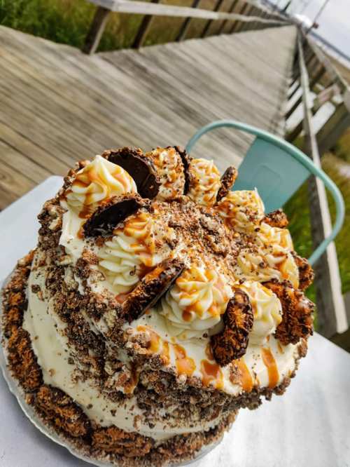 A layered cake topped with frosting, cookie pieces, and caramel drizzle, set against a wooden boardwalk backdrop.