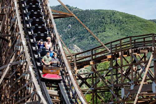A group of people enjoying a wooden roller coaster ride, descending a steep track with green mountains in the background.