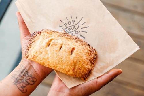 A hand holding a golden-brown pastry on a branded paper bag from Pop Pie Co.