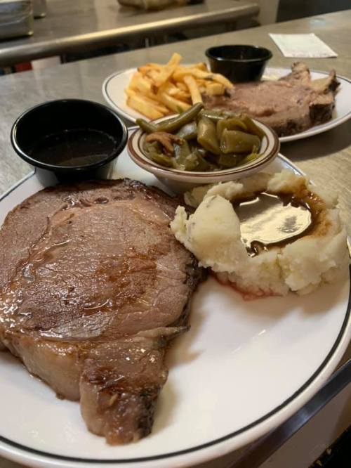 A plate featuring a juicy prime rib, mashed potatoes with gravy, green beans, and fries, with dipping sauces on the side.