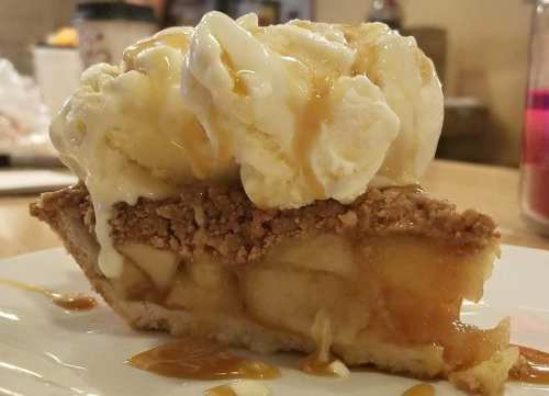 A slice of apple pie topped with vanilla ice cream and drizzled with caramel sauce on a white plate.