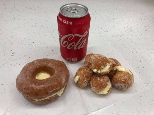 A glazed donut filled with cream next to a can of Coca-Cola and several cream-filled donut holes on a napkin.