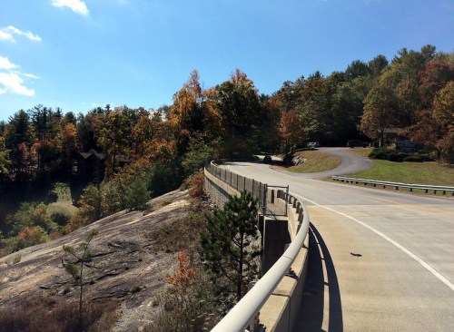 A winding road curves through a colorful autumn landscape with trees and a clear blue sky.
