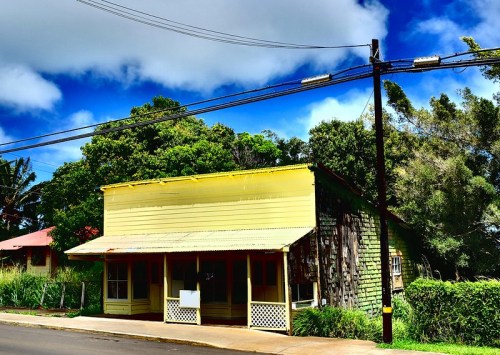 A yellow, rustic building with a corrugated roof, surrounded by greenery and under a blue sky with clouds.
