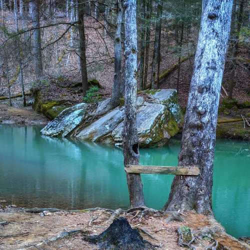 A serene forest scene featuring a turquoise river, large rocks, and trees with a wooden signpost nearby.