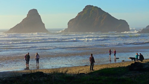 People walking along a beach with two large rock formations in the background and gentle waves lapping at the shore.