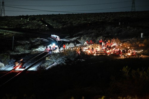 A nighttime scene of a gathering near train tracks, with people and lights illuminating the area amidst a desert landscape.