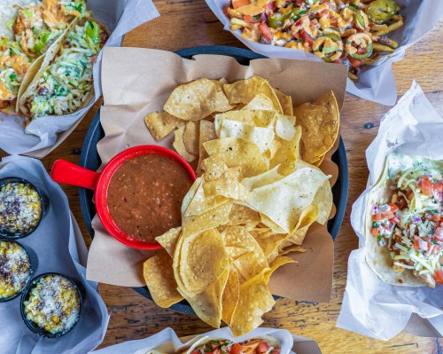 A platter of tortilla chips with salsa, surrounded by various taco dishes and toppings on a wooden table.