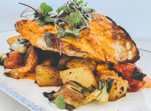 A plated dish featuring a golden-brown fish fillet topped with microgreens, served with roasted potatoes and vegetables.