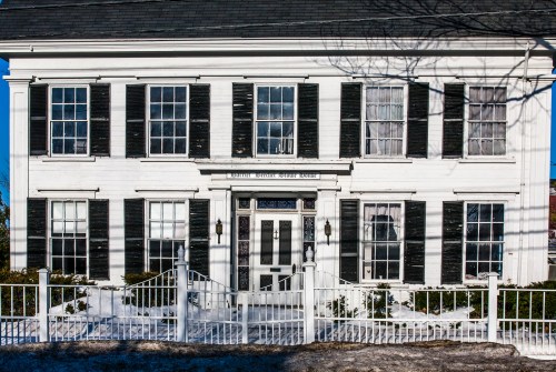 A historic white house with black shutters, featuring a fenced yard and a clear blue sky in the background.