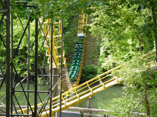A yellow roller coaster twists through lush green trees above a calm body of water.