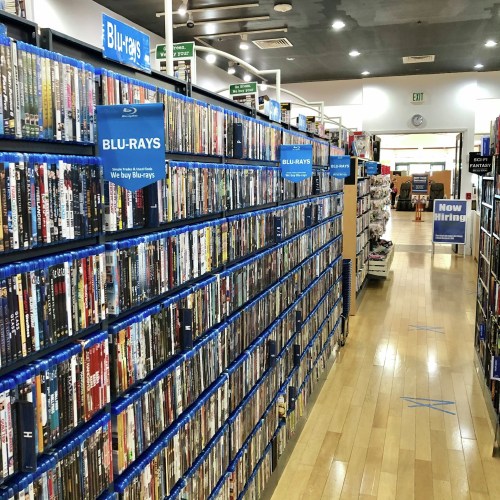 A store aisle lined with shelves of Blu-ray movies, with a "Now Hiring" sign visible in the background.